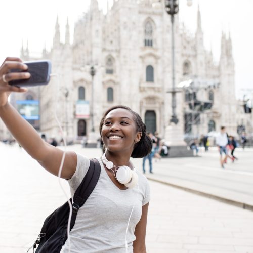 Young black woman taking selfie with smart phone hand hold with Milan cathedral in background, smiling - happiness, traveler, technology concept