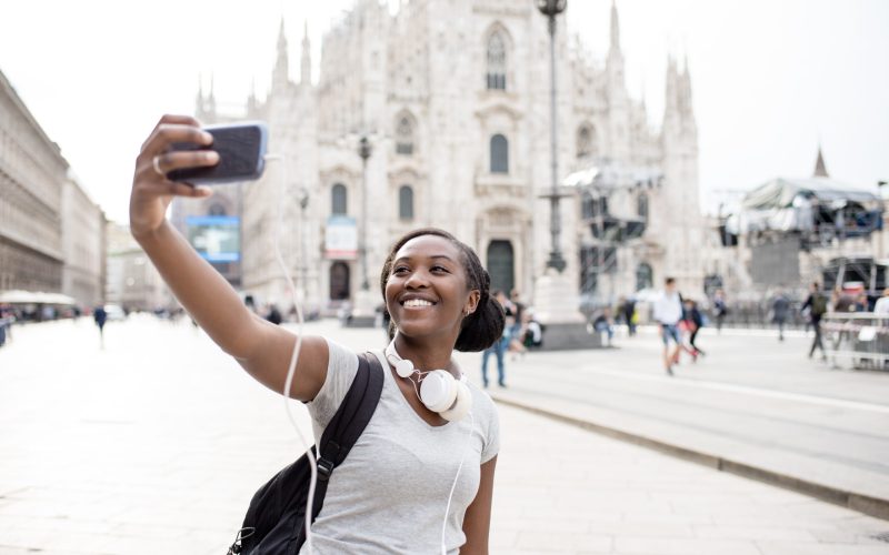 Young black woman taking selfie with smart phone hand hold with Milan cathedral in background, smiling - happiness, traveler, technology concept